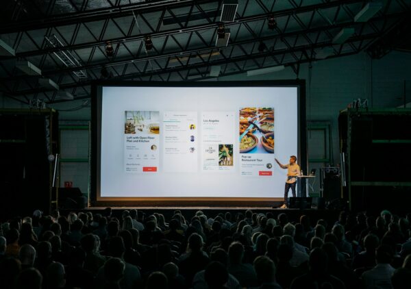 person discussing while standing in front of a large screen in front of people inside dim-lighted room
